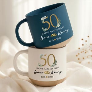 May include: Two ceramic mugs, one navy blue and one cream, stacked. Each mug features a gold glitter "50" with floral accents, the words "Happy Anniversary", and the names "Laura & Kenny" with the years "1975 2025".