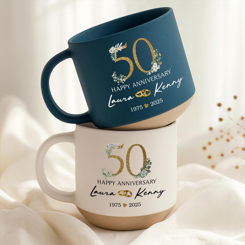 May include: Two ceramic mugs, one navy blue and one cream, stacked. Each mug features a gold glitter "50" with floral accents, the words "Happy Anniversary", and the names "Laura & Kenny" with the years "1975 2025".