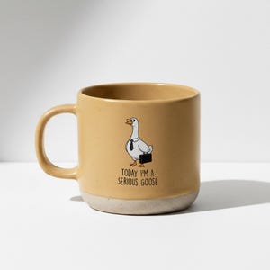 Può includere: Una tazza in ceramica beige con base bianca. La tazza presenta un'oca dei cartoni animati con cravatta e valigetta, con la scritta "TODAY I'M A SERIOUS GOOSE". La tazza ha un manico grande.