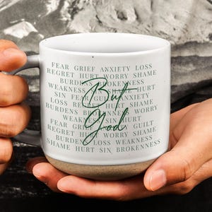 May include: A white ceramic mug with a gray handle and a tan base. The mug features the words "But God" in a cursive font, surrounded by words like "fear," "grief," and "shame."