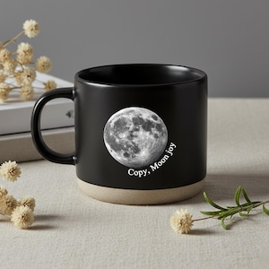 Peut inclure: Mug en céramique noire avec un motif de pleine lune et le texte "Copy, Moon joy". Le mug a une base beige et une anse noire. Le mug est posé sur une surface neutre avec des fleurs séchées.