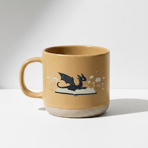 May include: Mustard-coloured ceramic mug featuring a black dragon illustration on an open book. The dragon has detailed wings and is surrounded by white flowers. The mug has a speckled finish and a light beige base.