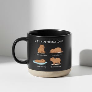 May include: Black ceramic mug with a beige base, featuring the text "DAILY AFFIRMATIONS" and illustrations of capybaras. Affirmations include "I am confident," "I feel relaxed," "I am strong," and "I am worthy."
