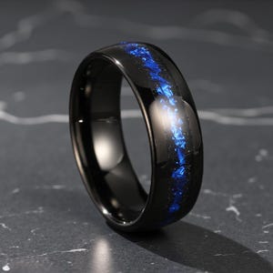 Könnte beinhalten: Ein schwarzer Ring mit glänzender Oberfläche. Der Ring hat eine leuchtend blaue Einlage, die durch die Mitte verläuft. Der Ring ist vor einem dunklen, strukturierten Hintergrund platziert, was sein elegantes Design hervorhebt.