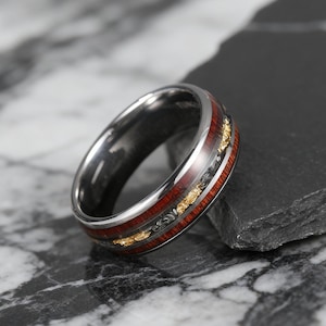 Peut inclure: Gros plan d'une bague argentée avec une incrustation en bois rouge foncé et des accents dorés. La bague présente une finition polie et est mise en valeur sur un fond sombre. Design moderne et élégant.