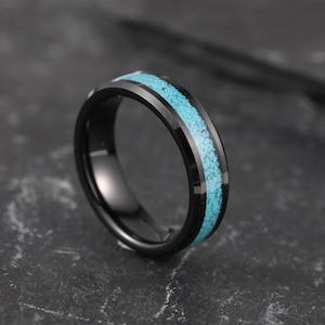 Peut inclure: Une bague noire avec une incrustation turquoise. La bague a une surface polie et réfléchissante avec une bande de pierres bleu-vert vif serties au centre. La bague est présentée sur une surface sombre et texturée.