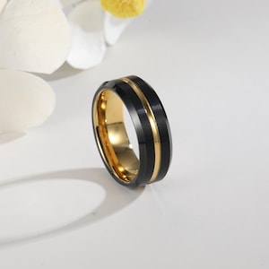 Puede incluir: Un anillo negro y dorado con un acabado pulido. El anillo presenta una banda negra con un centro dorado y una banda interior dorada. El anillo está sobre un fondo blanco con una flor.