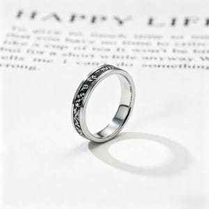 Könnte beinhalten: Ein silberner Ring mit einem detaillierten schwarzen Muster um das Band. Der Ring befindet sich vor einem weißen Hintergrund mit verschwommenem Text im Hintergrund. Der Ring ist ein einfacher Bandstil mit poliertem Finish.