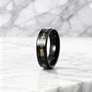May include: A black ring with gold leaf accents. The ring has a glossy finish and a band that is approximately 0.3 inches wide. The gold leaf is scattered throughout the black material, creating a unique pattern. The ring is set against a marble background.