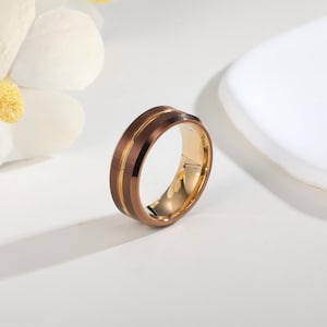 Puede incluir: Un anillo marrón y dorado con un acabado cepillado. El anillo presenta una banda central marrón flanqueada por dos bandas doradas. El interior del anillo también es dorado. El anillo está sobre un fondo blanco.