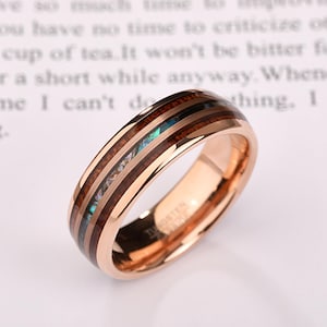 Peut inclure: Bague en tungstène or rose avec incrustation de bois et accents nacrés irisés. La bague présente une finition polie et une bande avec des incrustations alternées de bois foncé et d'ormeau. Le texte "TUNGSTEN" est visible à l'intérieur.