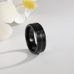 Può includere: Un anello nero in tungsteno con una fascia centrale scanalata. L'anello ha una finitura lucida e un bordo smussato. L'anello è una taglia 9.