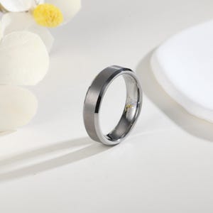 Könnte beinhalten: Ein silberfarbener Ring mit einem gebürsteten grauen Mittelband und polierten Kanten. Der Ring hat ein abgeschrägtes Design und eine glatte, metallische Oberfläche. Der Ring ist ein Schmuckstück.
