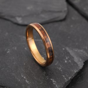 Può includere: Un anello color oro con un intarsio in legno marrone scuro. L'anello ha una finitura liscia e lucida e un design a fascia classico. L'intarsio in legno aggiunge un elemento naturale e strutturato all'estetica generale dell'anello.