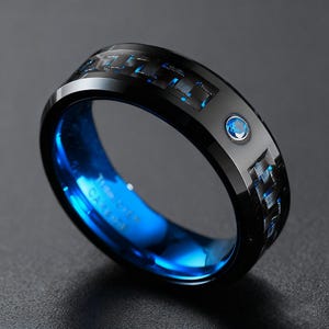 May include: A black ring with a blue interior and a blue gemstone. The ring has a carbon fiber weave pattern with blue accents. The ring is a close-up shot on a dark background.