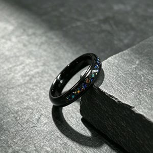 Peut inclure: Une bague noire dotée d'un anneau lisse et poli avec une incrustation centrale de fragments iridescents multicolores broyés. L'incrustation présente de petits morceaux réfléchissants dans des tons de bleu, vert et orange sur le métal.