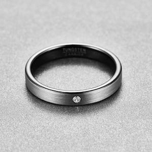 Puede incluir: Un anillo de carburo de tungsteno plateado con una pequeña gema transparente. El anillo tiene un acabado de metal cepillado y las palabras "TUNGSTEN CARBIDE" están grabadas en el interior.