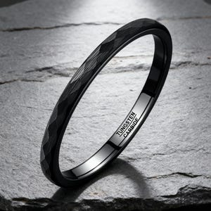 Peut inclure: Une bague noire en carbure de tungstène avec une surface texturée. L'intérieur de la bague est poli et porte l'inscription "TUNGSTEN CARBIDE". La bague est présentée sur une surface en pierre grise.