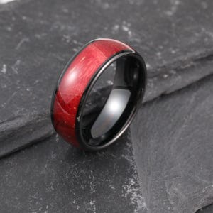 Puede incluir: Un anillo negro con una incrustación de madera roja. El anillo tiene un acabado pulido y la veta de la madera es visible. El anillo está sobre un fondo oscuro y texturizado. Un primer plano resalta los detalles del anillo.