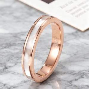 Può includere: Anello in oro rosa con intarsio bianco centrale. L'anello ha una finitura lucida e un design semplice ed elegante. La fascia è larga circa 0,6 cm.