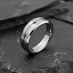 Peut inclure: Une bague argentée avec une bande centrale brossée et des bords polis. La bague porte l'inscription gravée "Love Can" à l'intérieur. La bague est présentée sur une surface sombre et texturée.