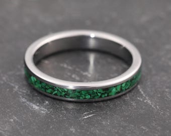 Könnte beinhalten: Ein silberner Ring mit einem Band aus leuchtend grünen, zerkleinerten Steinen. Der Ring ist kreisförmig und liegt auf einer dunklen, strukturierten Oberfläche. Das Design des Rings ist schlicht, aber auffällig.