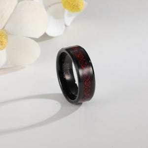 May include: Black tungsten carbide ring with a red Celtic knot design. The ring has the words "Tungsten Carbide" engraved on the side. The ring is a band style and is likely for a wedding or fashion accessory.