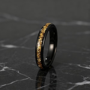 Peut inclure: Une bague noire avec une bande d'éclats d'or. La bague est posée sur un fond sombre, soulignant le contraste entre la bande noire et les éclats d'or. La bague est de conception simple.