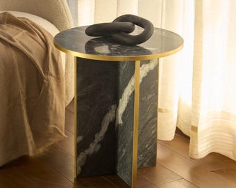 May include: A small, round side table with a black marble top and gold trim. The base is a dark green marble with gold accents. A dark gray decorative chain sits on top of the table. An open book rests on the wooden floor.