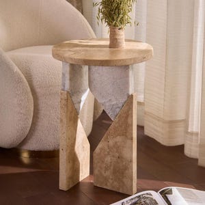 Op de afbeelding: Een moderne bijzettafel met een ronde, lichtgekleurde bovenkant en een geometrische basis. De basis is gemaakt van lichtbeige steen. Een kleine plant in een geweven pot staat op de tafel. Een open boek ligt op de vloer.