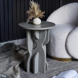 May include: A gray side table with a round top and a unique, sculptural base. A small white vase with dried flowers sits on the table. The table is next to a white armchair and a patterned rug.