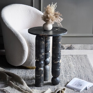 May include: A small, round side table with a black speckled top and three cylindrical legs. The legs are dark with a speckled pattern. A white, curved armchair and a grey rug are in the background. A small vase with dried flowers sits on the table.