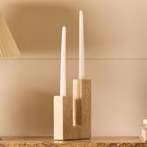 May include: A beige stone candle holder with a U-shaped design featuring two vertical columns of different heights. Two slender white taper candles are inserted into the top of each column. The stone has a textured, porous surface finish.