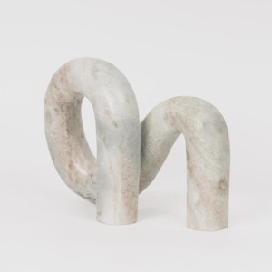 Op de afbeelding: Een sculpturaal object vervaardigd uit gepolijste steen met een gladde, continu gebogen vorm. Het materiaal vertoont een gemarmerd patroon met zachte grijze, witte en subtiele roze-beige aders door de afgeronde, buisvormige structuur.