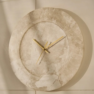 May include: A circular wall clock featuring a beige and cream marbled stone face with a smooth, polished texture. The clock has three thin, metallic gold-toned hands positioned at the center, with no numbers or markings on the clock face.
