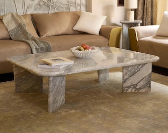May include: A rectangular marble coffee table with a gray and white veined pattern. The table has a smooth surface and is supported by four rectangular legs. A decorative bowl with fruit and a book are on the table. The table is in a living room setting.
