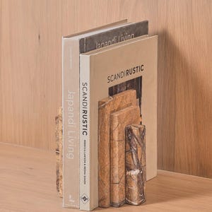 May include: Two rectangular bookends crafted from brown marble with visible veining, supporting two books titled Japandi Living and SCANDIRUSTIC. The bookends feature a tiered, stepped design with polished surfaces and natural stone patterns in shades of tan and dark brown.