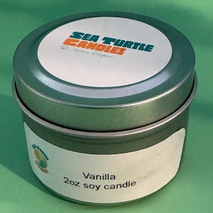 May include: A small, round, green metal container with a white label that reads "Vanilla 2oz soy candle." The lid has a white circle with the words "SEA TURTLE CANDLES" in blue and orange. The candle sits on a green surface.