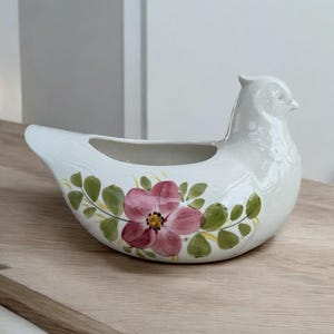 Peut inclure: Pot de fleurs en céramique blanche en forme d'oiseau avec une fleur rose et un motif de feuilles vertes. Le pot a une zone creuse pour les plantes et une tête d'oiseau avec un petit œil rose. Sur une surface en bois.