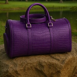 May include: A vibrant purple barrel-shaped duffel bag featuring a textured crocodile skin pattern. The bag includes two top handles, a detachable shoulder strap, and gold-toned metal hardware accents on the sides and handle attachments.