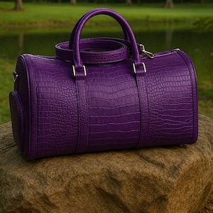Könnte beinhalten: Eine leuchtend violette, zylinderförmige Reisetasche mit einer strukturierten Krokodilleder-Optik. Die Tasche verfügt über zwei Tragegriffe, einen abnehmbaren Schulterriemen und goldfarbene Metallbeschläge an den Seiten und Griffbefestigungen.