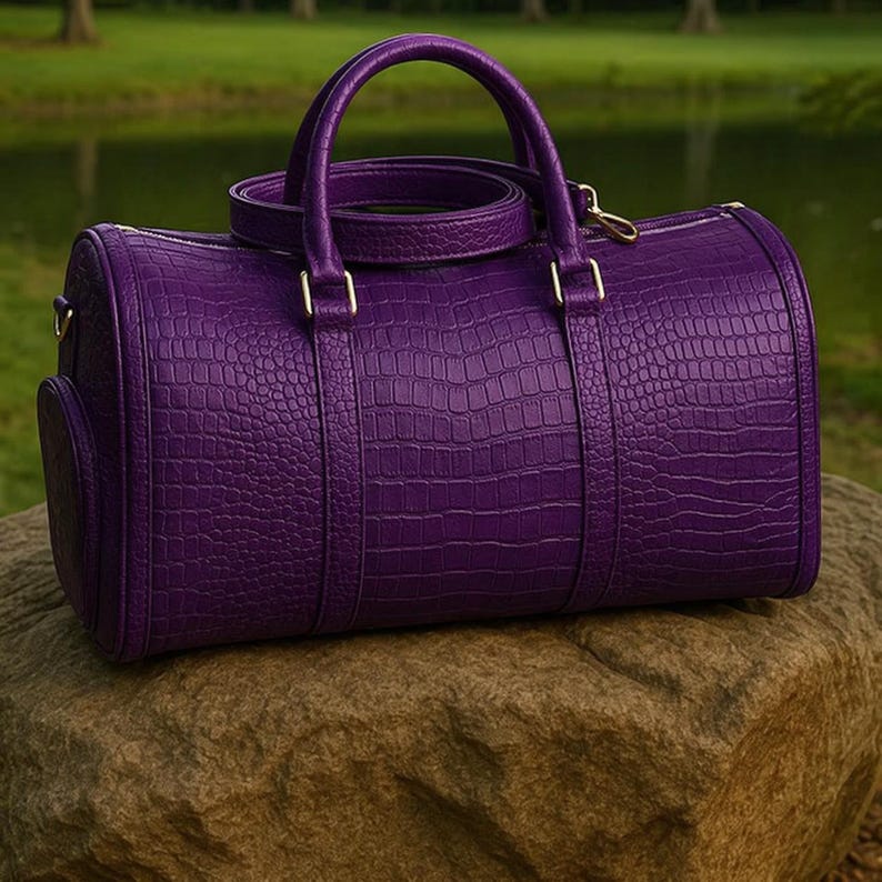 May include: A vibrant purple barrel-shaped duffel bag featuring a textured crocodile skin pattern. The bag includes two top handles, a detachable shoulder strap, and gold-toned metal hardware accents on the sides and handle attachments.