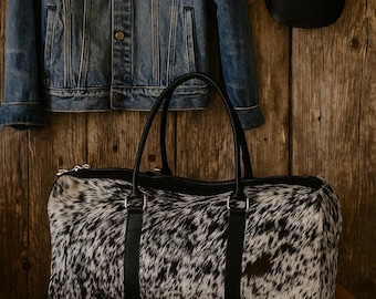 May include: A black and white cowhide duffel bag with black leather straps and handles sits on a mustard-colored quilted surface. A denim jacket and a black and white patterned baseball cap hang on a wooden wall.