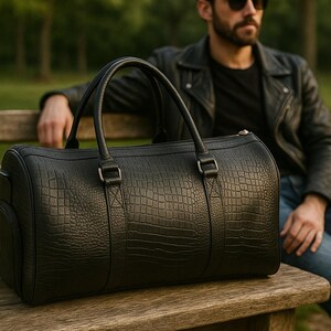 May include: A black duffel bag with a textured crocodile skin pattern, featuring two sturdy top handles, silver-toned metal hardware, and a cylindrical shape. The bag is constructed with reinforced stitching at the handle attachments.