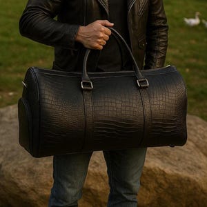 May include: A black duffel bag with a textured crocodile skin pattern, featuring two sturdy top handles, silver-toned metal hardware, and a cylindrical shape. The bag is constructed with reinforced straps that extend down the front and back panels.