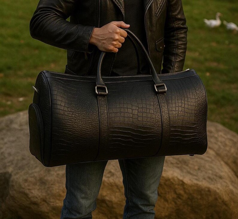 May include: A black duffel bag with a textured crocodile skin pattern, featuring two sturdy top handles, silver-toned metal hardware, and a cylindrical shape. The bag is constructed with reinforced straps that extend down the front and back panels.