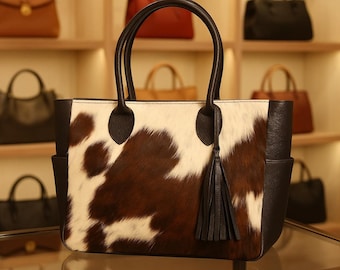 May include: A rectangular tote bag with a cowhide pattern in brown and white. The bag has black leather handles and trim, with a matching tassel. Other handbags are visible on shelves in the background.