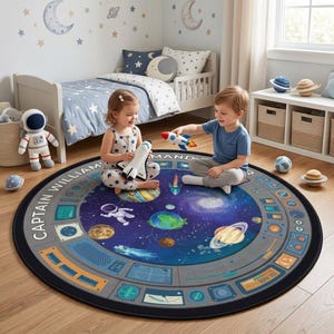 Peut inclure: Tapis rond sur le thème de l'espace, avec un design bleu foncé et gris. Il présente des planètes, des étoiles et un graphisme de centre de commande. Le texte "CAPTAIN WILLIAM" est imprimé sur le bord. Deux enfants jouent avec des fusées et une navette spatiale jouet.