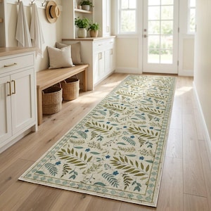 May include: A long rectangular runner rug featuring a cream-colored base with a repeating botanical pattern of green fern leaves and small blue flowers, finished with a decorative geometric border along the edges of the fabric floor covering.