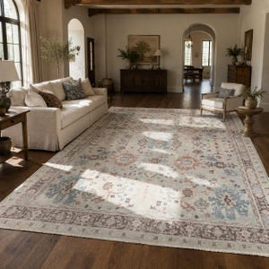 Puede incluir: Una alfombra rectangular grande con un patrón floral y de medallones intrincado y desteñido en tonos apagados de azul, terracota y crema. La alfombra tiene un borde ancho de color marrón oscuro con un motivo geométrico y flecos cortos.